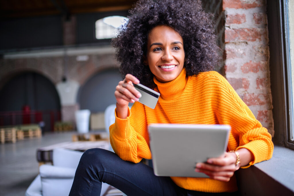 Photo of woman sitting by window smiling with a credit card and tablet